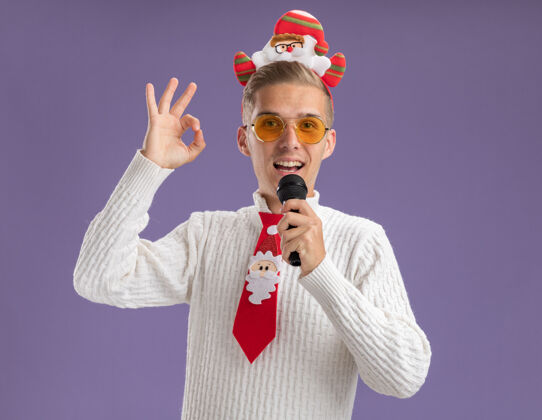 眼镜印象深刻的年轻帅哥戴着圣诞老人的头带和领带戴着眼镜拿着麦克风近嘴看着相机做ok标志隔离在紫色背景上领带小伙子抱着