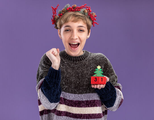 紫色快乐的年轻漂亮女孩戴着圣诞花环拿着圣诞树玩具和日期看着相机做是的手势孤立在紫色背景上快乐日期花圈