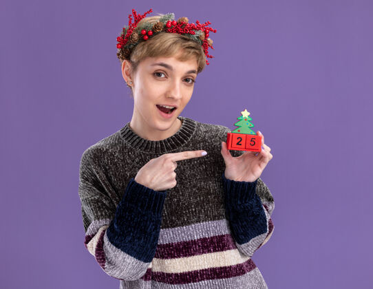 年轻给人印象深刻的年轻漂亮女孩戴着圣诞花环拿着圣诞树玩具和日期指着它看着隔离在紫色背景上的相机头花圈日期