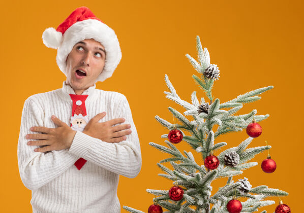 圣诞令人印象深刻的年轻帅哥戴着圣诞帽和圣诞老人领带站在装饰圣诞树附近双手交叉放在胸前看着橙色背景上孤立的一面壁板圣诞老人十字架