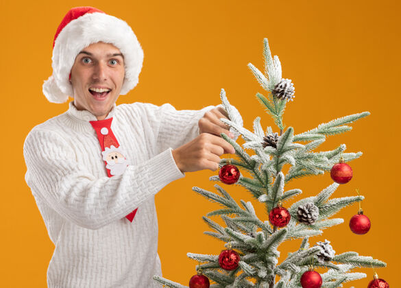印象令人印象深刻的年轻帅哥戴着圣诞帽 打着圣诞老人的领带站在圣诞树旁 用圣诞球装饰圣诞树 看着隔离在橙色背景上的相机橙色装饰品圣诞老人