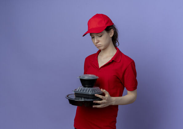 悲伤悲伤的年轻漂亮的送货女孩穿着红色制服 戴着帽子 看着紫色背景上的食品容器 还有复印空间红色女孩帽子