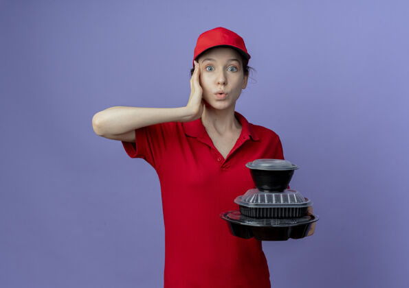 触摸令人印象深刻的年轻漂亮的送货女孩穿着红色制服和帽子举行食品容器触摸寺庙隔离紫色背景与复制空间帽子持有紫色
