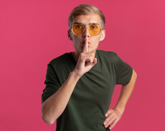 手势穿着绿色衬衫戴着眼镜的年轻帅哥表现出沉默的姿势把手放在臀部隔离在粉红色的墙上脸人男人