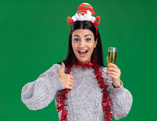 印象年轻的白人女孩戴着圣诞老人的头带 脖子上戴着金箔花环 手里拿着一杯香槟 看着相机 在绿色的背景上孤立地竖起大拇指圣诞老人圣诞快乐向上