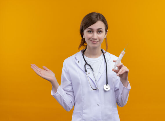 显示自信的年轻女医生穿着医用长袍和听诊器拿着注射器 在孤立的橙色空间里展示空手听诊器持有手