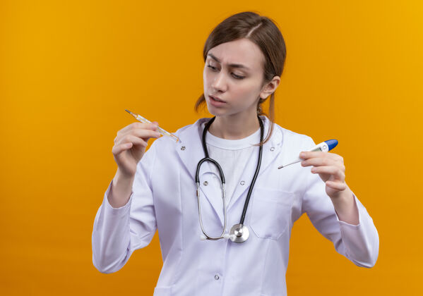 女性困惑的年轻女医生穿着医用长袍和听诊器 拿着温度计在孤立的橙色空间里看着他们长袍医生橙色
