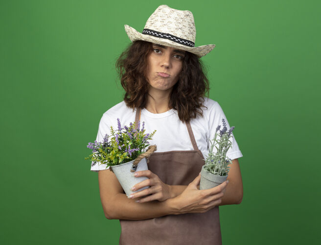 交叉穿着制服的悲伤的年轻女园丁戴着园艺帽在花盆里拿着花和交叉花制服园艺