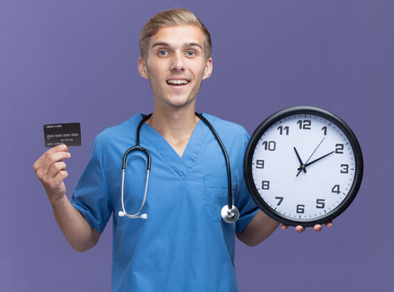 制服微笑的年轻男医生穿着医生制服 手持听诊器 拿着信用卡 墙上的挂钟被隔离在蓝色的墙上时钟信用医生