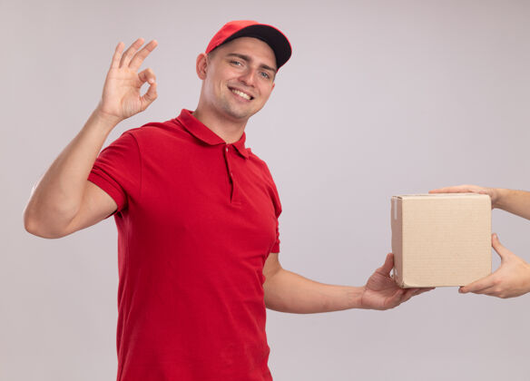 展示面带微笑的年轻送货员穿着制服 戴着帽子 在白色的墙上向客户展示了一个好的手势帽子给予送货