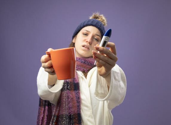 拿着未出嫁的生病少女 穿着白袍 戴着冬帽 戴着围巾 手里拿着一杯茶 温度计隔离在紫色上年轻的茶围巾