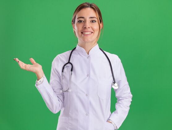 微笑面带微笑的年轻女医生穿着医用长袍 听诊器伸开的手被隔离在绿色的墙上听诊器长袍穿着