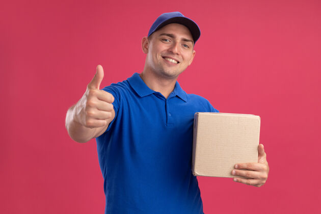 微笑面带微笑的年轻送货员穿着制服 戴着帽子的盒子 在粉红色的墙上孤立地竖起大拇指展示制服送货