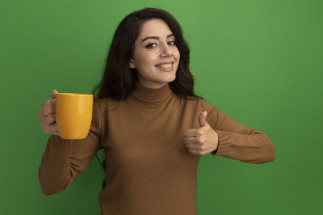 年轻微笑着的年轻漂亮的女孩端着一杯茶 在绿色的墙上孤立地竖起大拇指表演抱着杯子