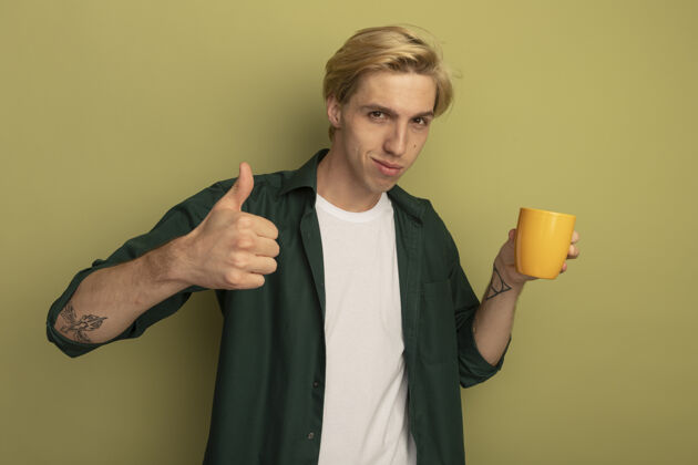 茶高兴的年轻金发小伙子穿着绿色t恤 端着一杯茶 竖起大拇指拇指穿抱着