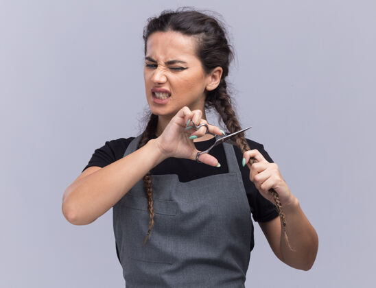剪愤怒的年轻女理发师穿着制服用剪刀剪头发隔离在白墙上剪刀制服年轻