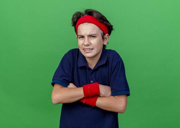 年轻皱眉的年轻帅气的运动男孩戴着头带和护腕 戴着牙套 站在封闭的姿势看前面 隔离在绿色的墙壁上 有复制空间腕带人市民