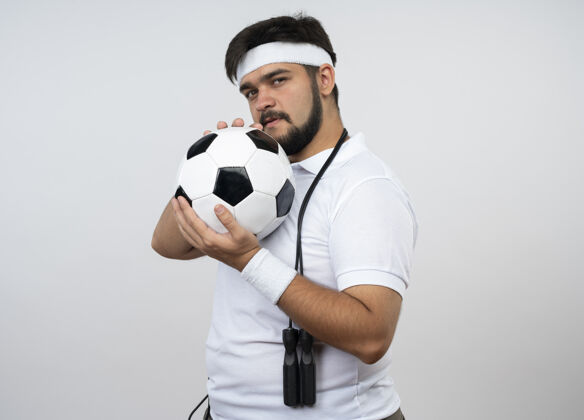 头带自信的年轻运动型男子戴着头带和腕带 肩上系着跳绳 把球绕在脸上 隔离在白色墙壁上 留有复制空间自信年轻抱着
