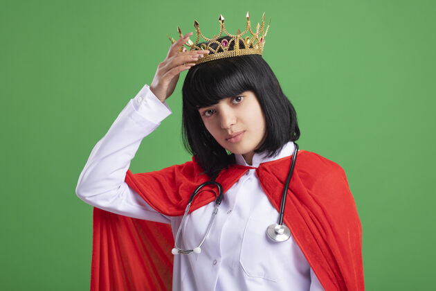 头自信的年轻超级英雄女孩戴着听诊器 穿着医用长袍和斗篷 头戴皇冠 与绿色隔离绿色信心医疗