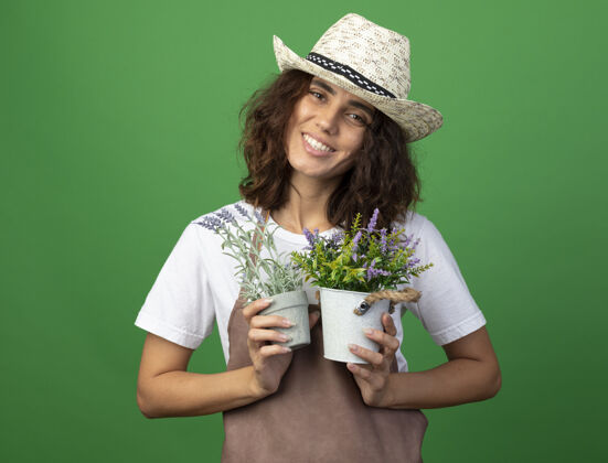 花微笑着的年轻女园丁 穿着制服 戴着园艺帽 把花盆里的花隔离在绿茵场上花盆园艺举行