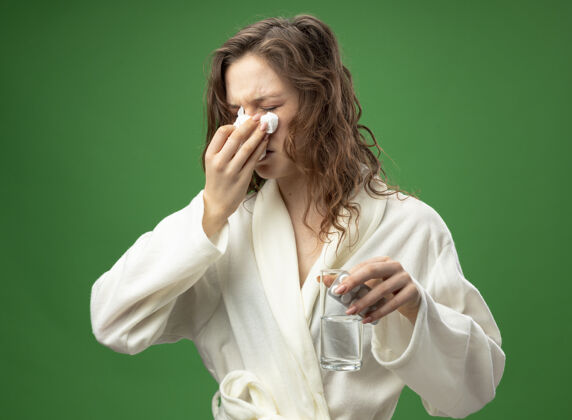 长袍年幼体弱多病的女孩闭着眼睛 穿着白色长袍 拿着一杯清水 用餐巾擦鼻子 隔离在绿色的地面上绿色鼻子擦