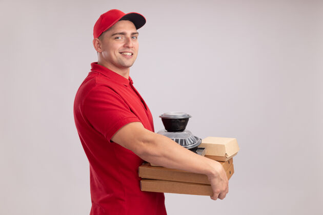 披萨面带微笑的年轻送货员穿着制服 戴着帽子 把食物容器放在隔离在白墙上的比萨饼盒上食物容器男人