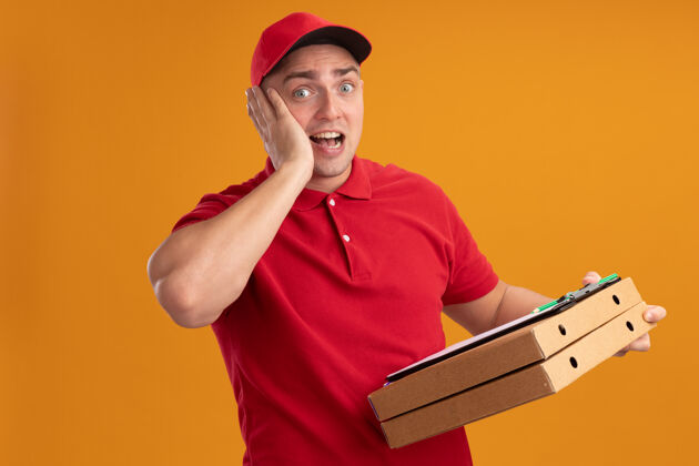 惊喜惊讶的年轻送货员穿着制服 戴着帽子 拿着披萨盒 手放在脸颊上 隔离在橙色的墙上拿着送货剪贴板