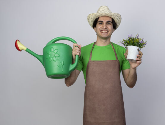 年轻人微笑着的年轻男园丁 穿着制服 戴着园艺帽 手里拿着花盆里的花和浇水罐男人戴着人