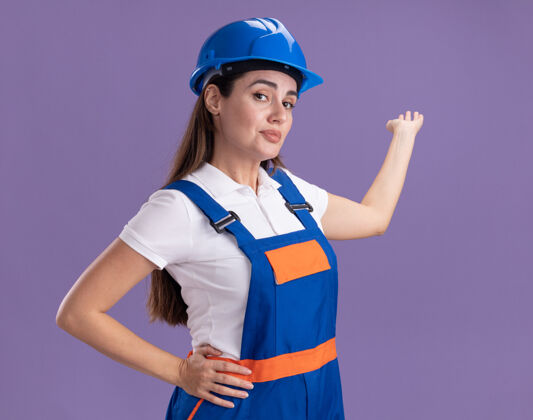 制服喜出望外的年轻女建筑工点着制服 用手在后面隔离着紫色的墙面上用复制的空间请年轻建筑工人