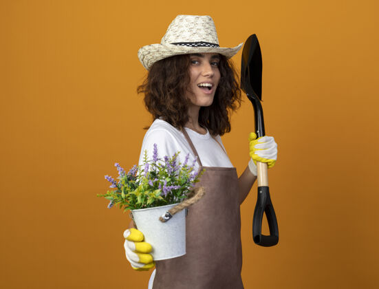 手套身着制服的年轻女园丁微笑着戴着园艺帽和手套 在花盆里捧着花花花园微笑