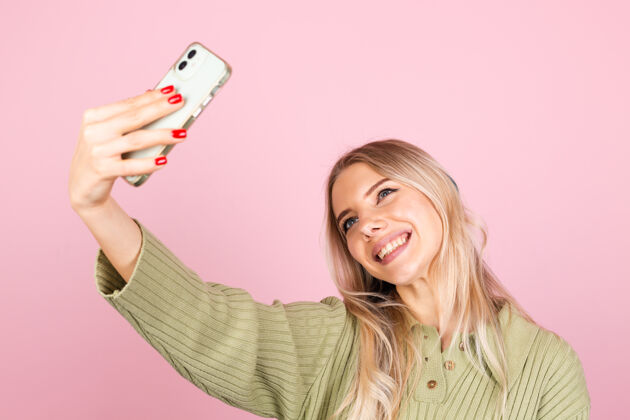 头发漂亮的欧洲女人在粉红色的墙上穿着休闲针织毛衣高兴脸手机