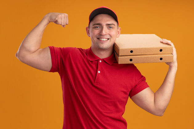 盒子微笑的年轻送货员身穿制服 头戴鸭舌帽 肩上扛着披萨盒 在橙色的墙上显示出强烈的手势制服微笑披萨