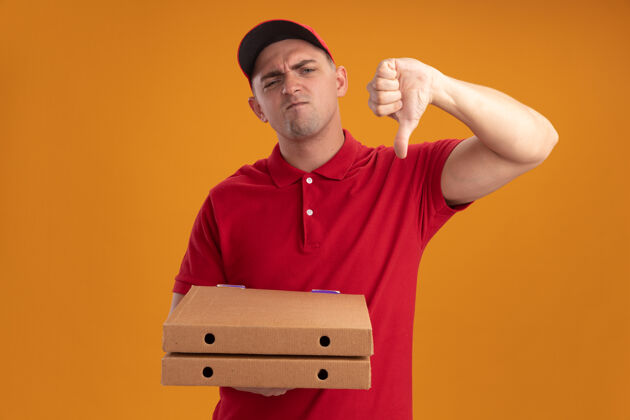 拿着年轻的送货员穿着制服 戴着帽子 拿着披萨盒 拇指朝下 被隔离在橙色的墙上披萨帽子送货