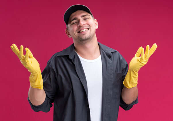 蔓延面带微笑的年轻汉姆森保洁员穿着t恤和帽子 戴着手套 双手分开放在粉红色的墙上微笑手套手