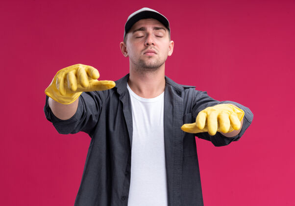 帽子闭着眼睛 年轻的汉姆森清洁家伙穿着t恤和帽子 戴着手套 伸出手孤立在粉红色的墙上清洁举行手套