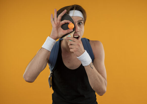 腕带快乐的年轻人 戴着背包 头上戴着头带 脸上戴着护腕 乒乓球拍和球被隔离在橙色的墙上乒乓球封面球