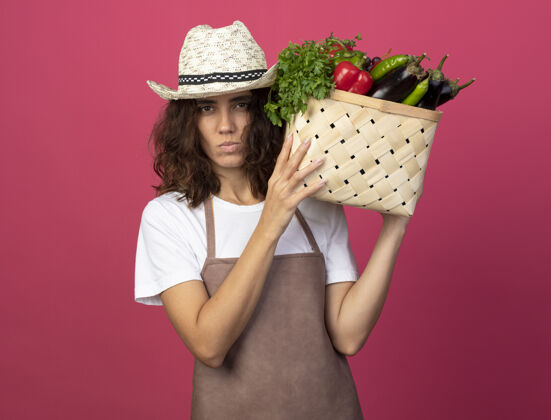 穿自信的年轻女园丁穿着制服戴着园艺帽把菜篮子放在肩上自信帽子女人