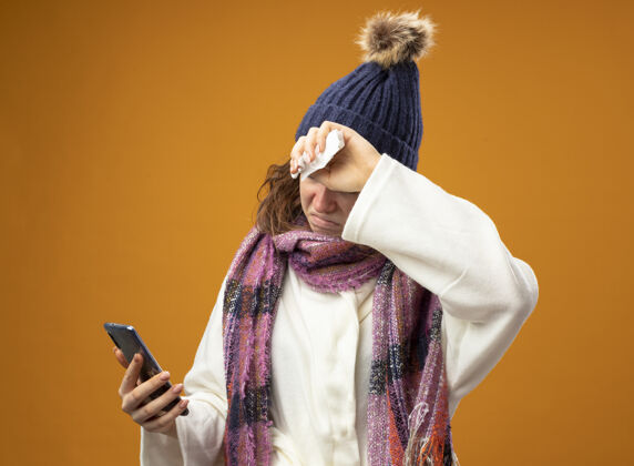 额头穿着白色长袍 戴着冬天的帽子 戴着围巾 手放在额头上 看着电话 孤立地站在橙色的墙上年轻长袍电话