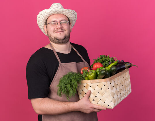 花园面带微笑的年轻男园丁戴着园艺帽 手里拿着蔬菜篮子 孤零零地站在粉红色的墙上穿男性持有