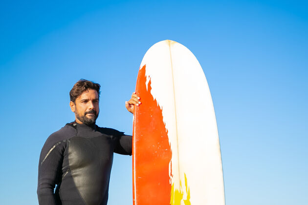 度假体贴的男性冲浪者站在冲浪板上的肖像穿着潜水服的黑发白种人拿着冲浪板向前看冲浪板极限海洋