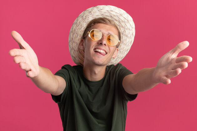 小伙子兴奋的年轻帅哥穿着绿色衬衫 戴着眼镜 戴着帽子 伸出双手孤立在粉红色的墙上帽子眼镜戴着