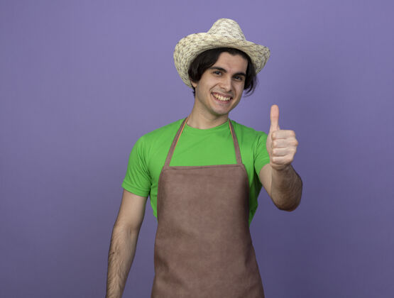 园艺微笑着的年轻男园丁穿着制服 戴着园艺帽 竖起大拇指年轻紫色花园