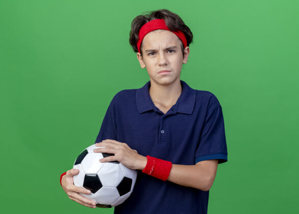 足球皱眉的年轻帅气的运动男孩戴着头带和护腕 戴着牙套 拿着足球 看着前面隔离在绿色墙上的复制空间头带脸抱着