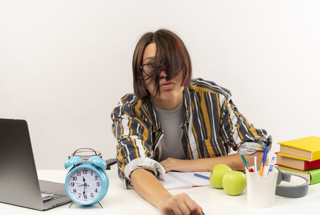 学生疲惫的年轻女生戴着眼镜坐在书桌旁用大学工具看着相机在白色背景上孤立年轻穿女孩