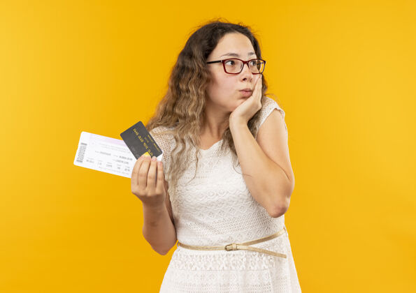 眼镜印象深刻的年轻漂亮的女学生戴着眼镜 背着包拿着票和信用卡 手放在脸上看着黄色隔离的一面票穿着信用