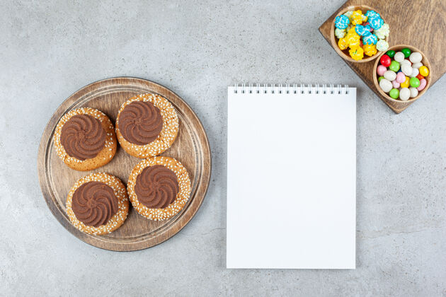 美味把饼干和两碗糖果堆在木板上 放在大理石背景的白色笔记本上高质量的照片糕点午餐笔记本