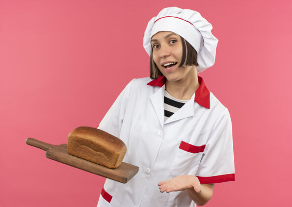 粉红身着厨师制服的快乐年轻女厨师手拿并指着插着粉红色面包的砧板喜悦持有女
