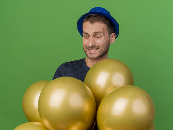 衣服失望的帅哥戴着蓝色的派对帽 抱着氦气球 看着隔离在绿色墙上的复制空间脸帽子失望
