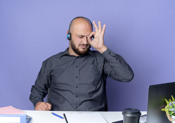 手势年轻秃顶的呼叫中心男子戴着耳机坐在办公桌旁 拿着工作工具做着看笔记本的手势 孤立在紫色背景上穿坐着秃头