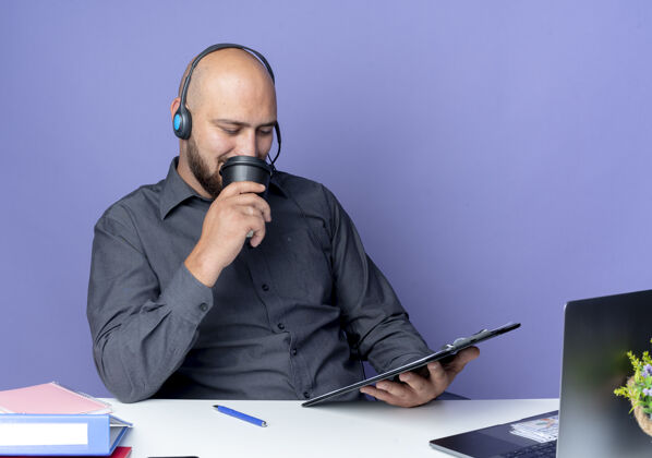 拿着年轻的秃头呼叫中心男子戴着耳机坐在办公桌旁 拿着工作工具 看着剪贴板 喝着紫色背景上孤立的咖啡穿剪贴板年轻